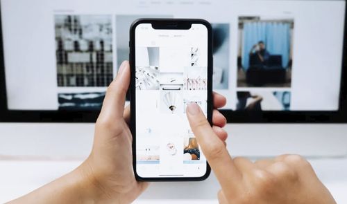 Primer plano de manos sosteniendo un smartphone frente a un monitor para localizar fotos en internet mediante una cuadrícula de imágenes.