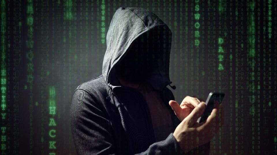 Una persona desconocida, con aspecto de hacker y encapuchada, usa un teléfono móvil con un fondo digital.