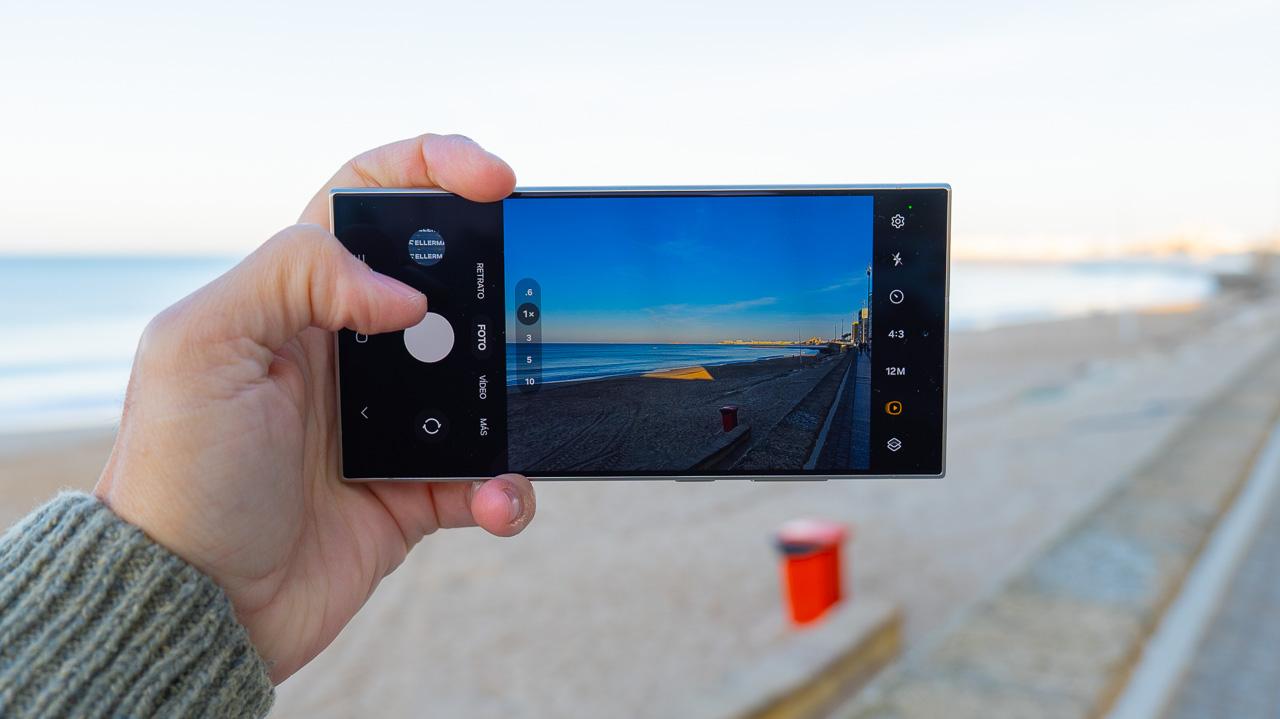 Mujer joven en el paseo de la playa sujetando en su mano un movil Galaxy S24 Ultra negro titanium utilizando la aplicación de fotos con el Zoom avanzado de hasta 10x de su smartphone Galaxy S24 Series. Utilizando las funciones de inteligencia artificial (AI).