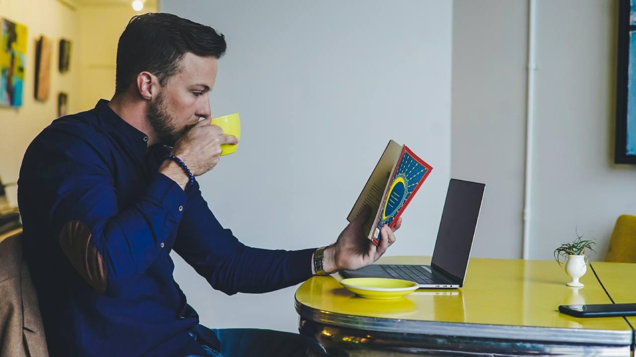 Un hombre lee un libro delante de su ordenador.