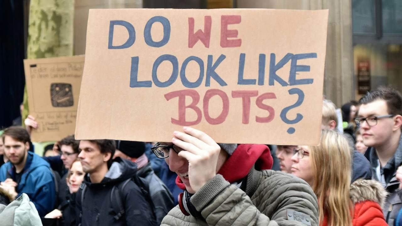 Un hombre lleva una pancarta que dice: "Do we look like bots?".