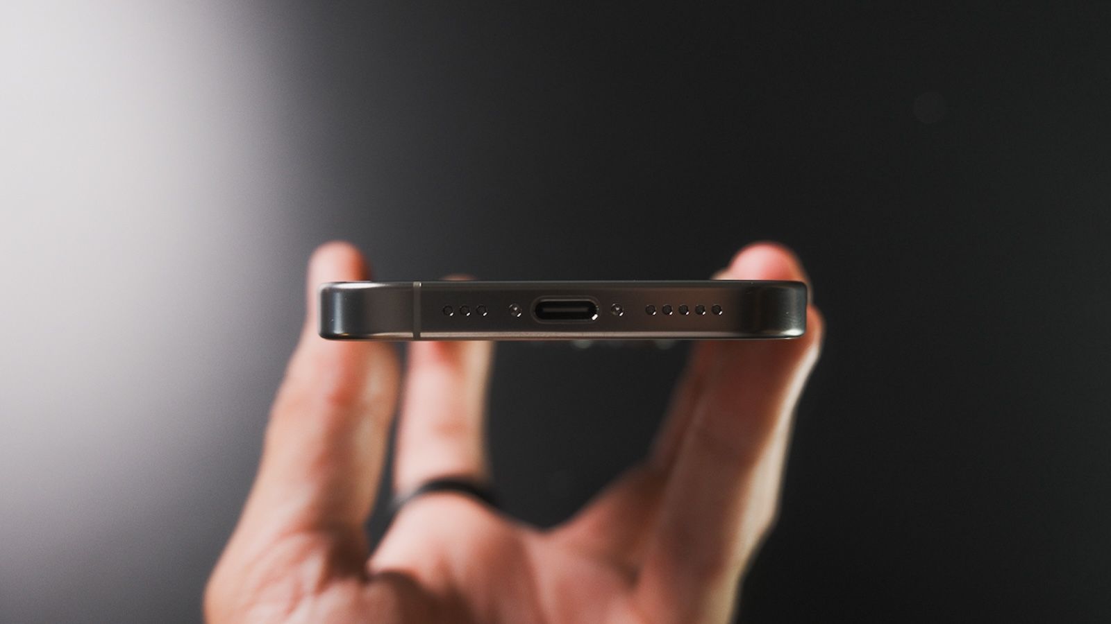 Vista detallada de la parte inferior de un iPhone 16 Pro color titanio, mostrando el puerto de carga USB-C en el centro y las rejillas de altavoz y micrófono a cada lado