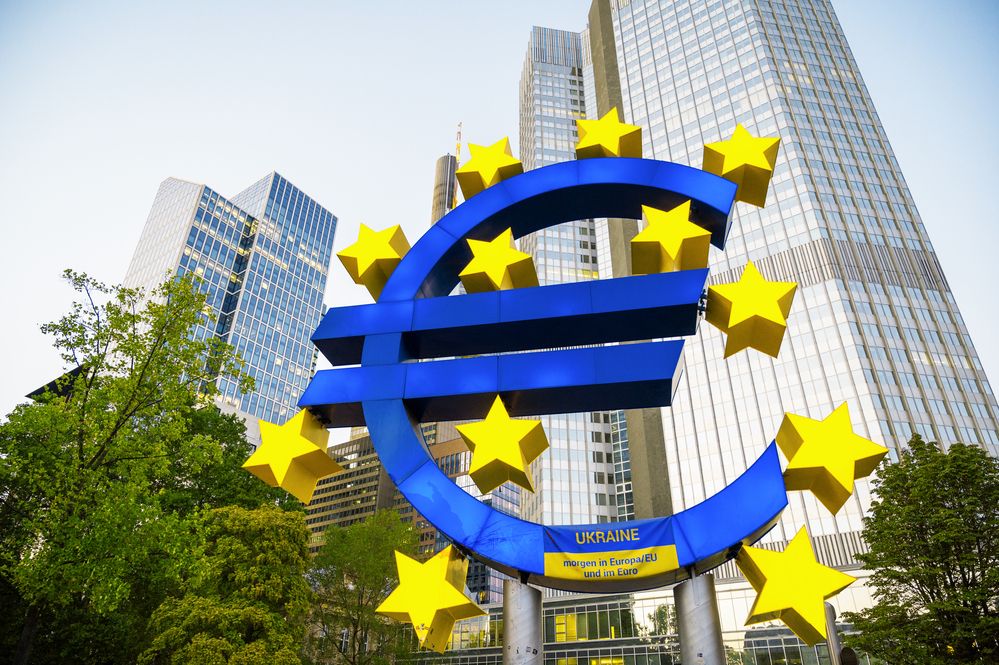 Gran escultura del símbolo del euro en color azul rodeada de estrellas amarillas frente a rascacielos en Fráncfort.