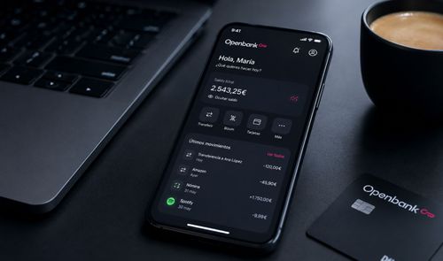 Smartphone mostrando la aplicación móvil de Openbank junto a un ordenador portátil, una taza de café y una tarjeta bancaria sobre una mesa oscura.