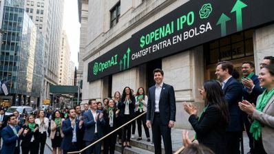 Sam Altman junto a un grupo de personas frente a la fachada del New York Stock Exchange con un cartel digital que anuncia la entrada en bolsa de OpenAI.