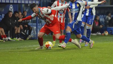 Dos futbolistas disputan el balón en un partido de la Primera Federación con el banquillo de fondo.