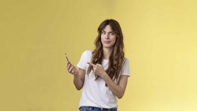 Mujer joven con camiseta blanca señalando sorprendida un teléfono móvil sobre un fondo amarillo.