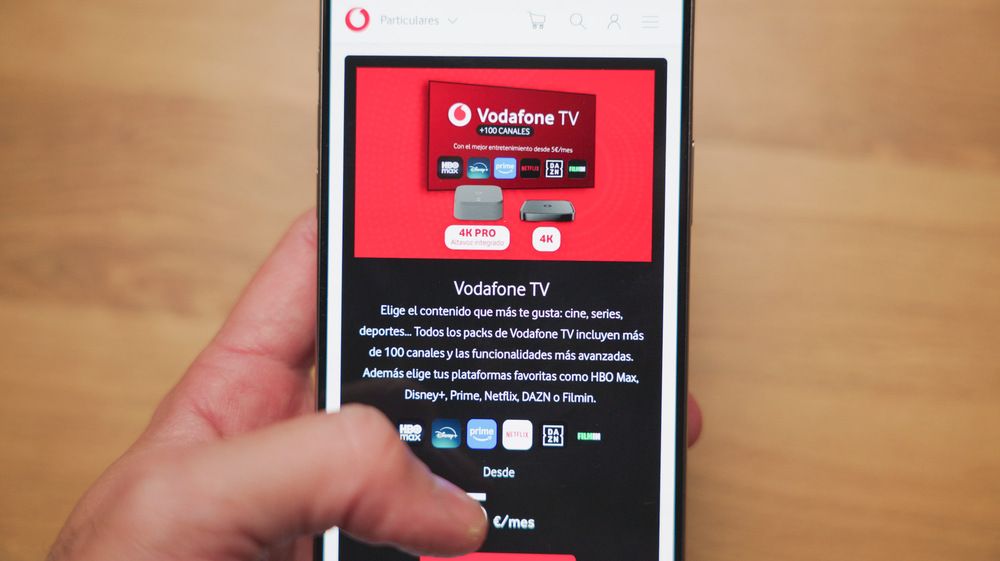 Primer plano de una mano sosteniendo un smartphone que muestra la oferta de Vodafone TV con información sobre canales y dispositivos decodificadores 4K.