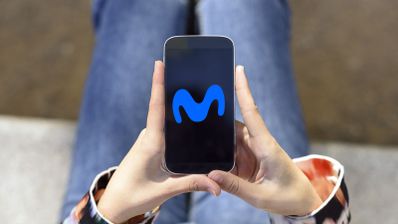 Primer plano de una persona sosteniendo un teléfono móvil con el logotipo azul de Movistar en la pantalla sobre un fondo desenfocado.
