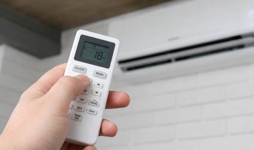 Mano sujetando un mando a distancia de aire acondicionado configurado a 18 grados frente a una unidad de split blanca en la pared.