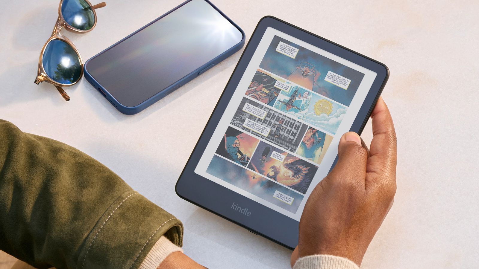 Persona sosteniendo un Kindle Colorsoft que muestra una página de cómic a color junto a un teléfono móvil y gafas de sol.