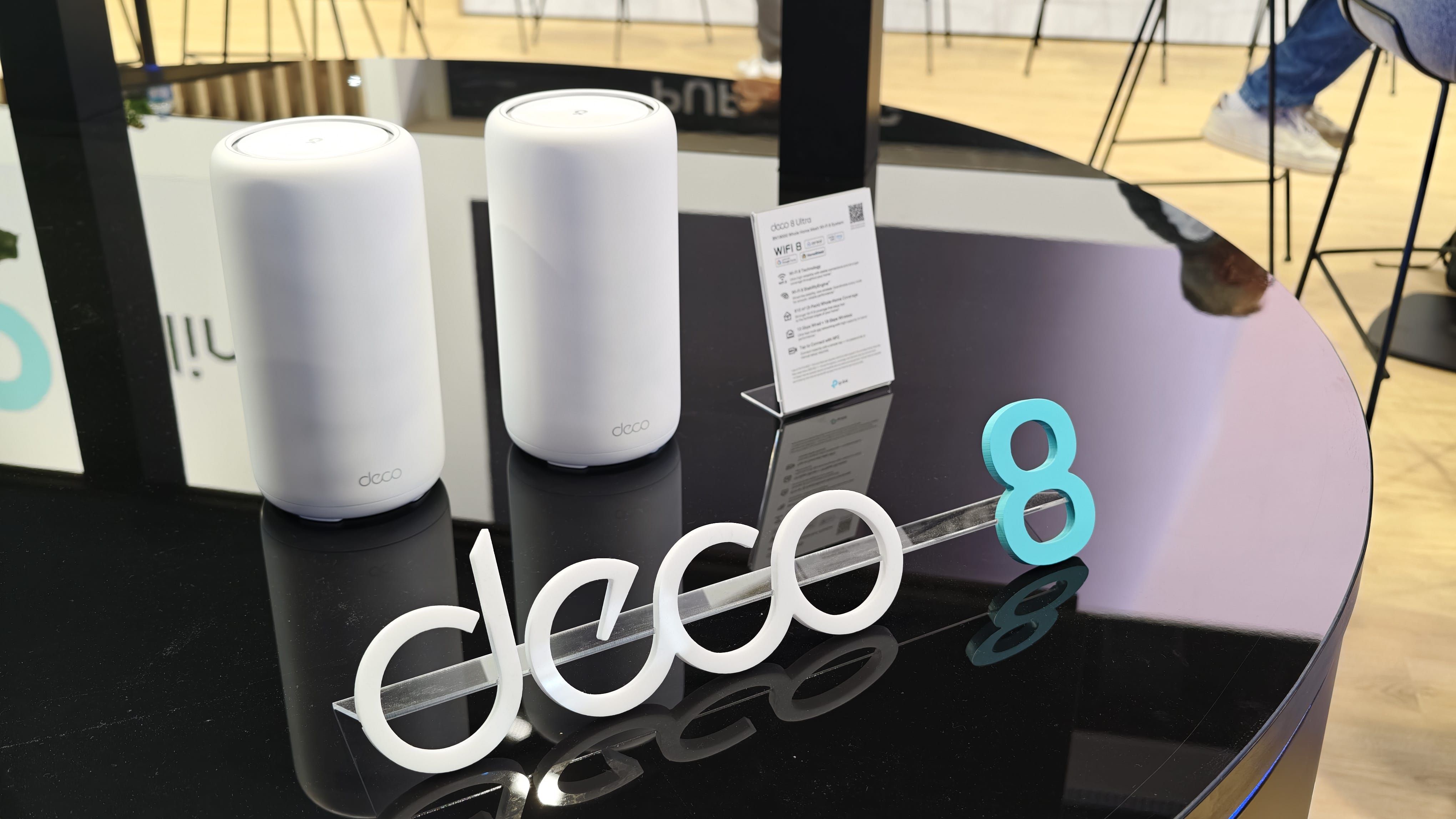 Dos nodos de red WiFi Mesh de color blanco cilíndrico junto a un logotipo físico que reza deco 8 sobre una mesa negra.