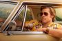 Brad Pitt conduciendo un coche clásico con gafas de sol y camisa amarilla estampada frente al letrero de Hollywood.