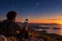 Persona sentada en una colina sosteniendo un telefono movil que muestra barras de cobertura satelital durante un atardecer, referencia 25T150640.