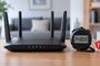 Router inalámbrico negro con cuatro antenas junto a un cronómetro digital que marca 24 horas y un icono de escudo de alerta sobre una mesa de madera.