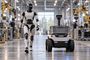 Un robot humanoide blanco y negro caminando junto a un vehículo autónomo de carga en una planta industrial.