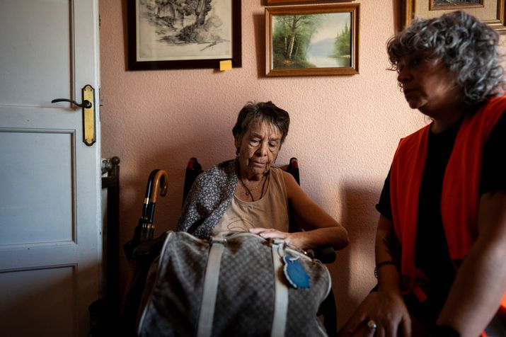 Mujer mayor sentada en el interior de una vivienda junto a una persona con chaleco naranja de asistencia y una bolsa de viaje.