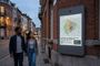 Pareja caminando por una calle empedrada junto a una pantalla informativa digital anclada en una fachada de ladrillo que muestra un mapa por zonas.