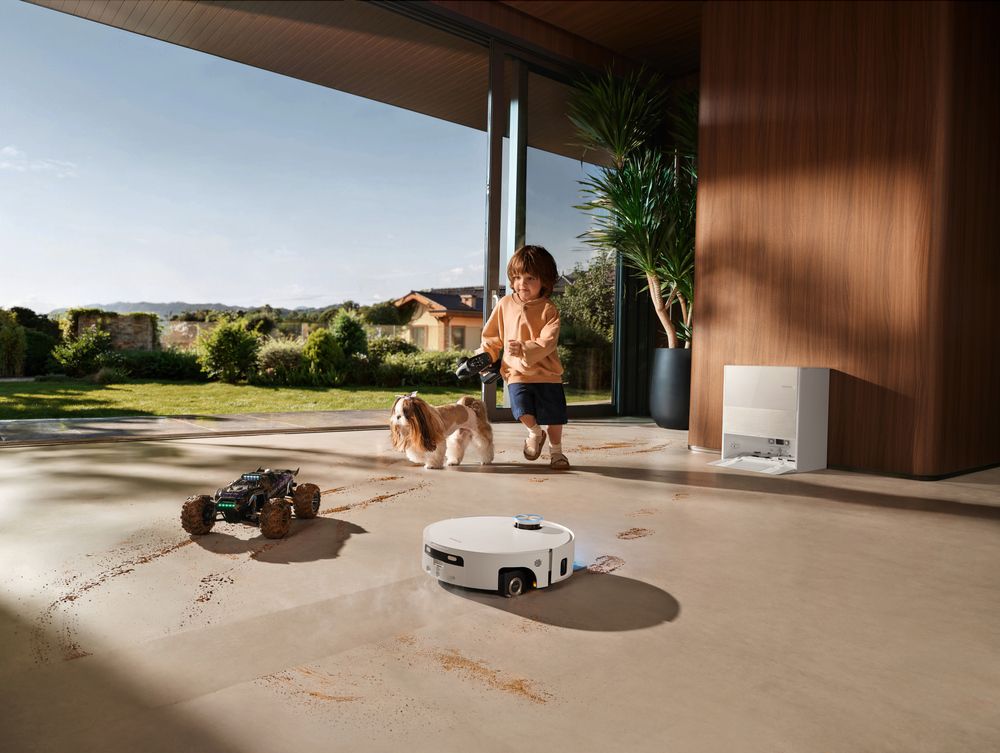 Robot aspirador blanco limpiando marcas de barro en el suelo de un salón mientras un niño y un perro juegan al fondo.