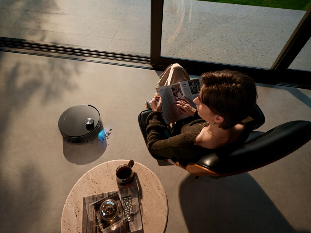 Vista cenital de una persona leyendo en un sillón mientras un robot aspirador limpia el suelo del salón.