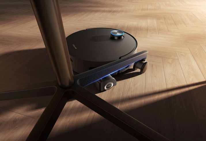 Robot aspirador de color negro circulando por un suelo de madera bajo la base de una silla de oficina.