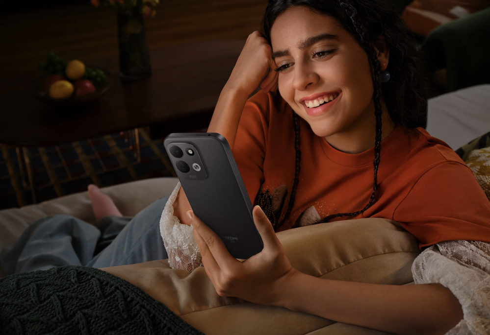 Mujer sonriente mirando la pantalla de un smartphone con doble cámara trasera en un entorno doméstico relajado.