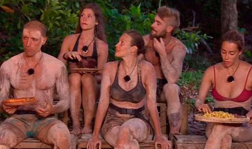 Grupo de concursantes de un programa de telerrealidad de supervivencia sentados en el suelo y cubiertos de barro mientras comen.