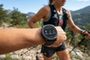 Primer plano de un reloj inteligente Garmin mostrando funciones de entrenamiento y asistencia en la muñeca de una deportista de montaña.