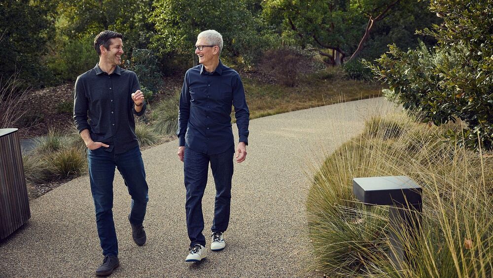 John Ternus y Tim Cook caminan juntos conversando en un sendero exterior rodeado de vegetación.