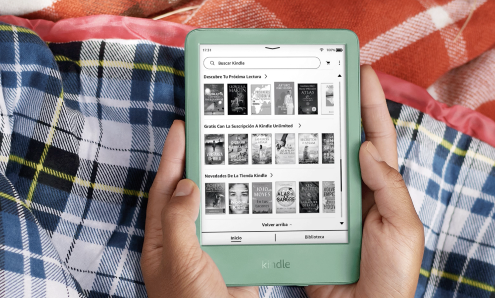 Manos sosteniendo un lector electrónico Kindle en color verde claro mostrando el catálogo de libros sobre una manta de cuadros.