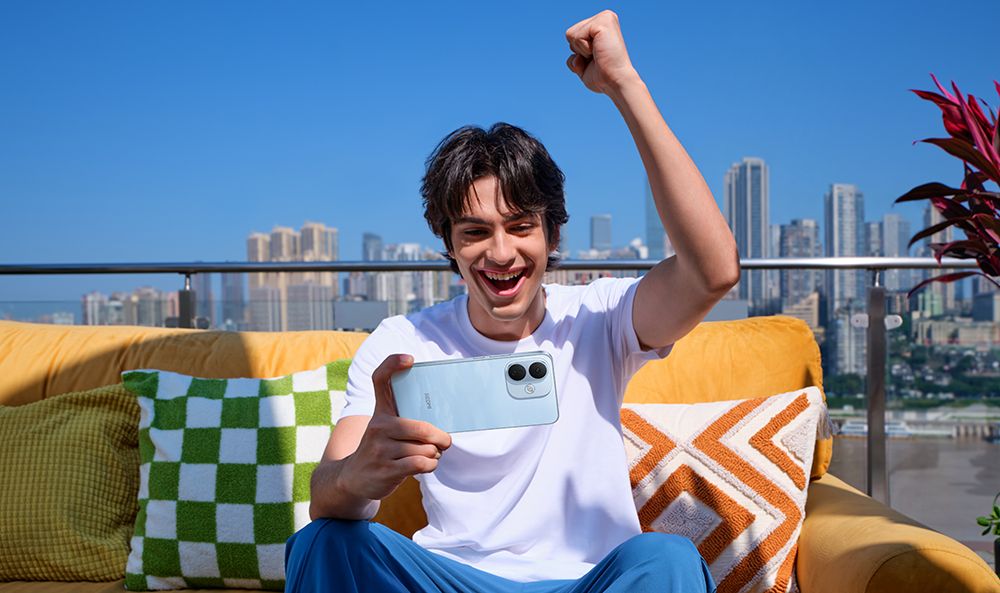 Joven celebrando con un Redmi Note 17 en la mano mientras está sentado en una terraza con rascacielos de fondo.