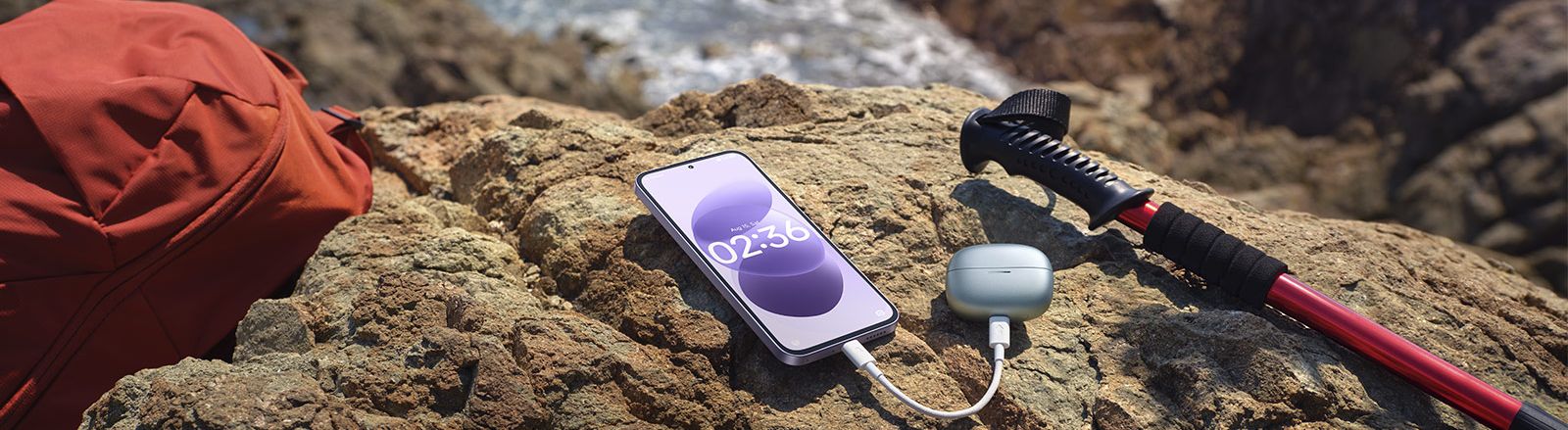 Teléfono Redmi Note 17 cargando unos auriculares inalámbricos sobre una roca en la costa.