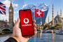 Mano sosteniendo un teléfono móvil con el logotipo de Vodafone frente a paisajes de Londres, Suiza y Turquía.