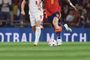 Primer plano de los pies de dos futbolistas durante un partido, uno con la equipación roja de la selección española y otro de blanco, disputando el balón sobre el césped.