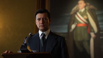 Hombre con traje frente a un micrófono en un atril con un retrato militar al fondo.