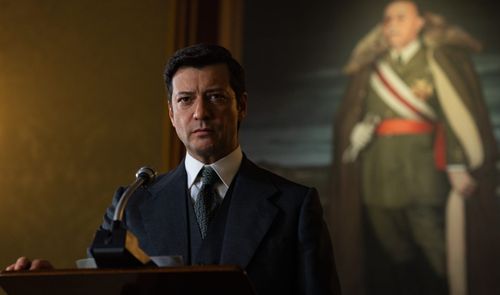 Hombre con traje frente a un micrófono en un atril con un retrato militar al fondo.
