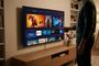 Persona utilizando un mando a distancia frente a una Smart TV colgada en la pared con menú de aplicaciones de streaming.