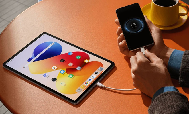 Tableta Redmi Pad 2 Pro cargando un teléfono móvil mediante conexión por cable sobre una mesa naranja.