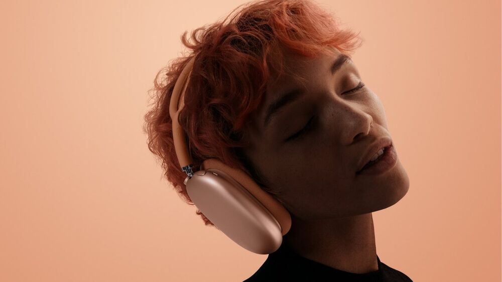 Primer plano lateral de una persona con auriculares AirPods Max 2 sobre las orejas y los ojos cerrados en un ambiente con tonos anaranjados.