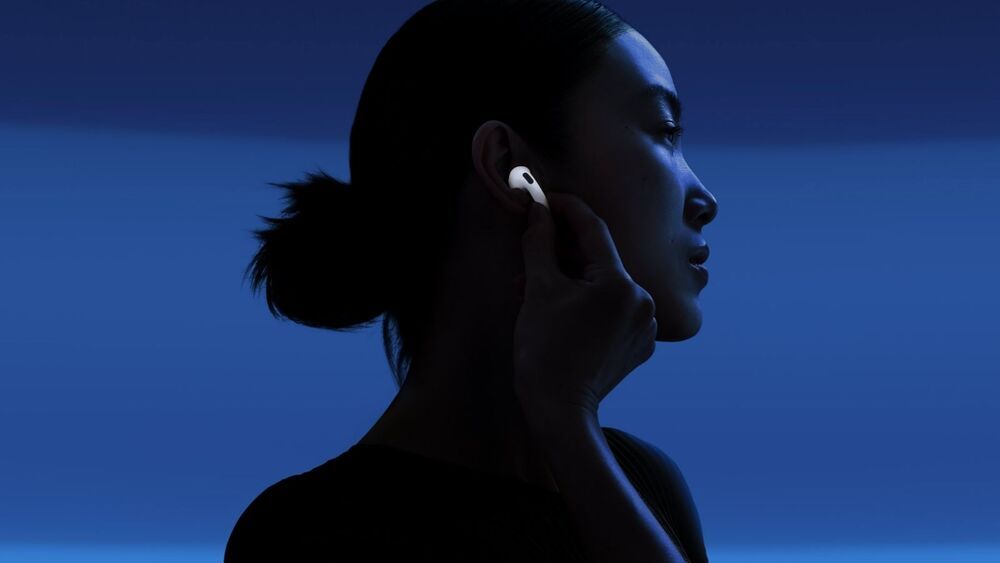 Mujer de perfil ajustándose un auricular inalámbrico blanco AirPods 5 bajo una iluminación azul tenue.