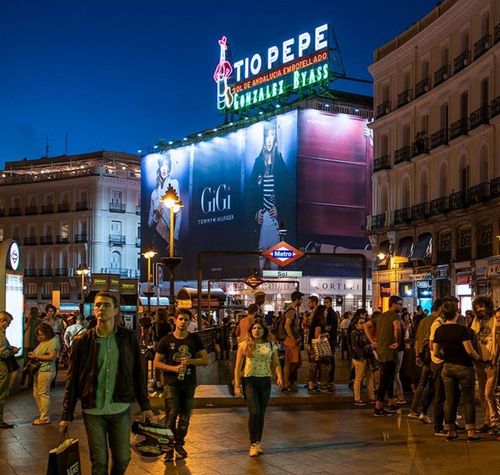 Personas caminando por la Puerta del Sol de Madrid al anochecer con el icónico cartel luminoso de Tío Pepe de fondo.