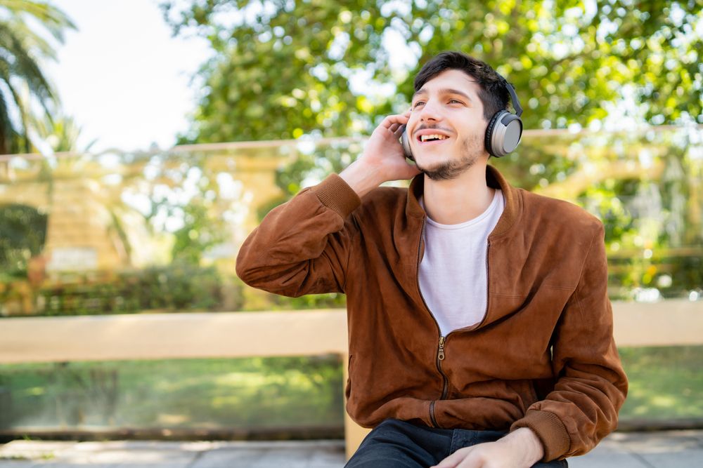 Hombre joven con chaqueta marrón escuchando música con auriculares de diadema en un parque.