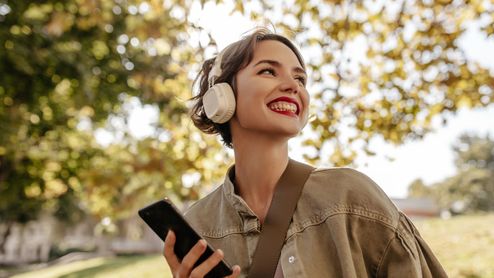 Mujer joven sonriendo mientras usa auriculares de diadema inalámbricos en un entorno exterior con árboles.