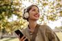 Mujer joven sonriendo mientras usa auriculares de diadema inalámbricos en un entorno exterior con árboles.