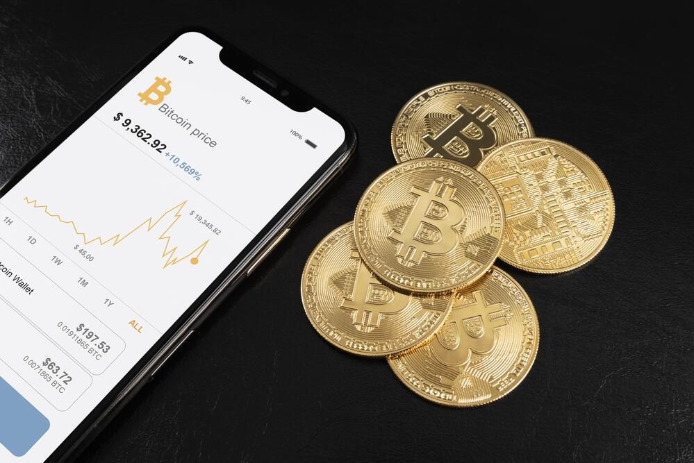 Smartphone con gráfica de cotización de Bitcoin junto a varias monedas físicas doradas de criptomonedas sobre fondo oscuro.