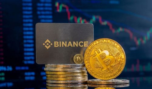 Tarjeta física de Binance junto a una moneda de Bitcoin sobre una pila de monedas con gráficas bursátiles al fondo.