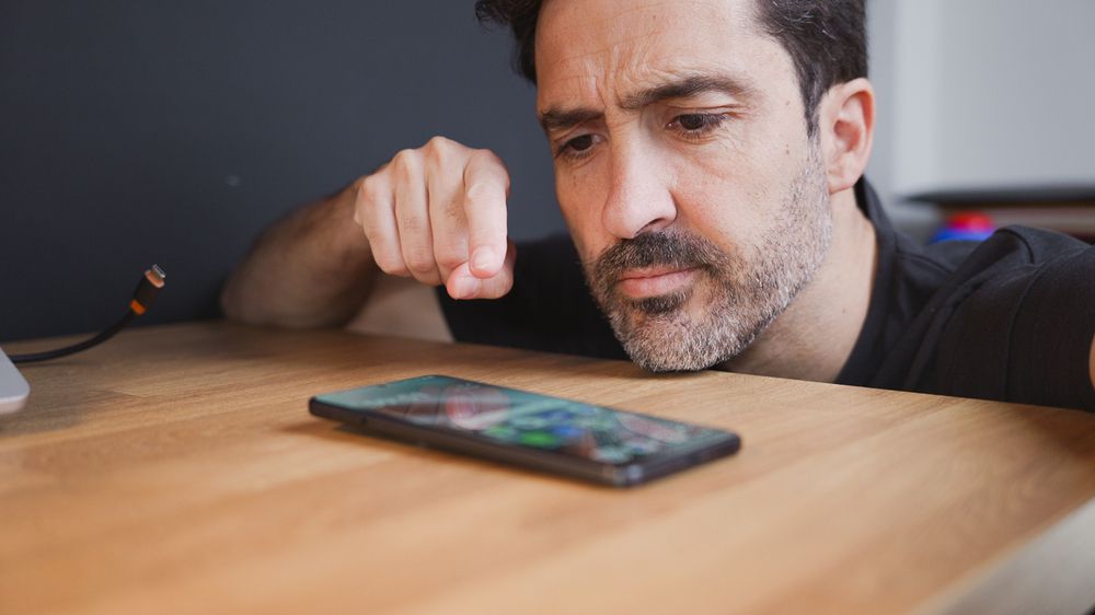 Hombre con expresión de duda mirando la pantalla de un teléfono Android apoyado en una mesa de madera.