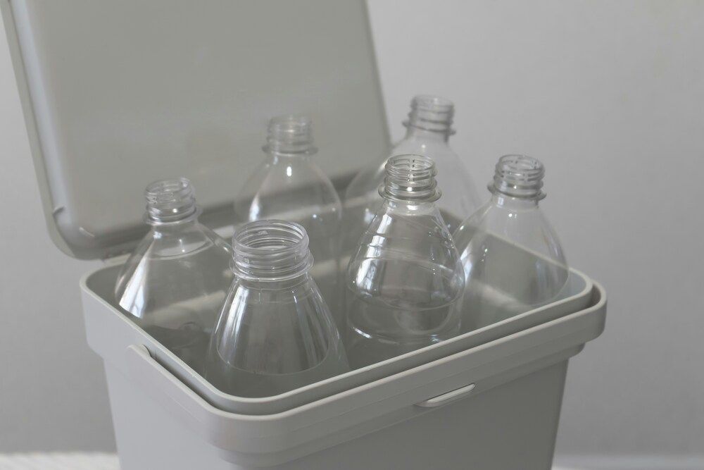 Botellas de plástico transparentes y vacías acumuladas dentro de un contenedor gris con la tapa abierta.