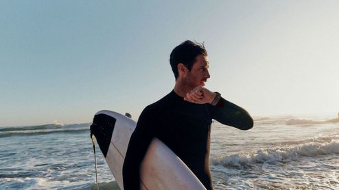 Surfista con traje de neopreno consultando su reloj inteligente fenix 9 mientras sujeta una tabla de surf en la orilla del mar.