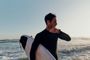 Surfista con traje de neopreno consultando su reloj inteligente fenix 9 mientras sujeta una tabla de surf en la orilla del mar.